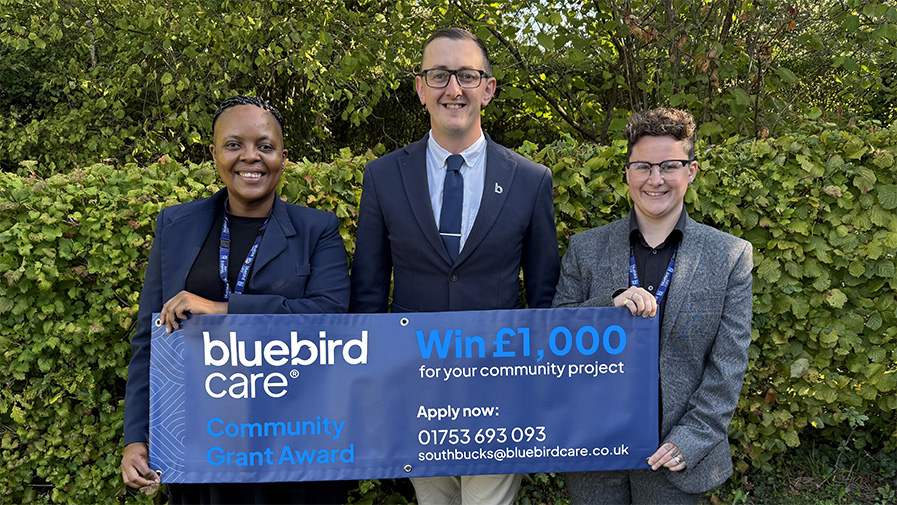 Bluebird Care staff stand in front of a banner which announces the launch of a £1,000 community grant, which will be awarded to fund a project that benefits the local area.